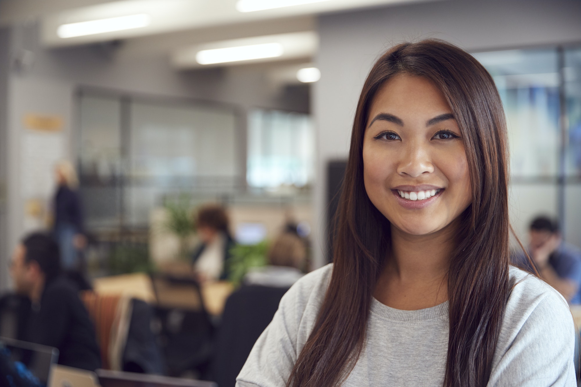 Head And Shoulders Portrait Of Smiling Young Asian Businesswoman Working In Busy Modern Office